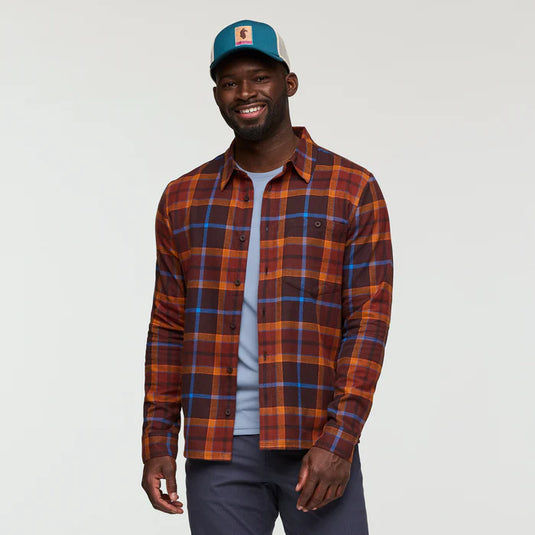 A man wearing a brown, red, and blue plaid flannel shirt with a collar, button-up style, and roll-up button cuffs.
