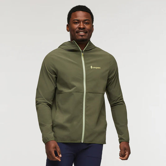A man wearing a green Cotopaxi Vuelta Performance Windbreaker jacket with a hood, featuring back and shoulder vents, and elastic binding on cuffs and hem.