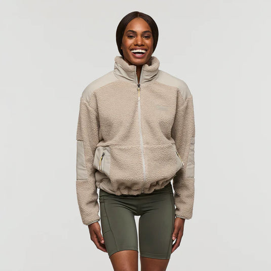 A woman wearing a beige Cotopaxi Bacano high-pile fleece jacket with a mandarin collar, zippered hand pockets, and retro-inspired patchwork.