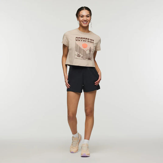 A woman standing, wearing black shorts with a visible wide waistband and a beige t-shirt with Cotopaxi text on it, suitable for summer casual wear.