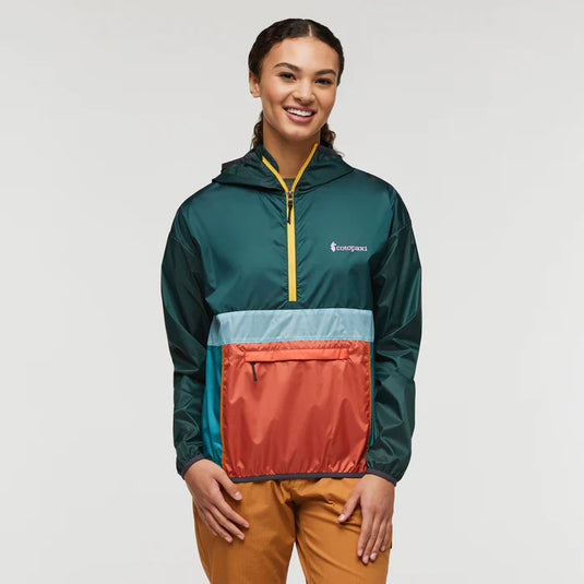 A woman wearing a green, blue, and orange half-zip windbreaker jacket with a Kangaroo hand pocket, and yellow zippered front pocket, paired with brown pants.