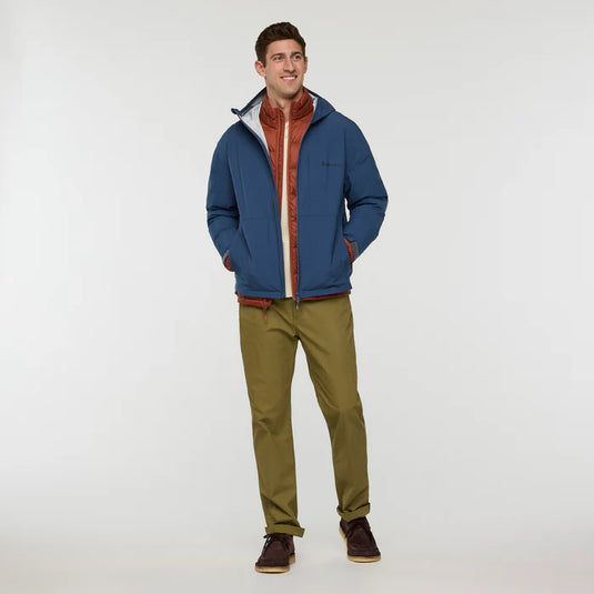 Man wearing a blue cotopaxi zip rain jacket with a red interior, green pants, and brown shoes on a white background