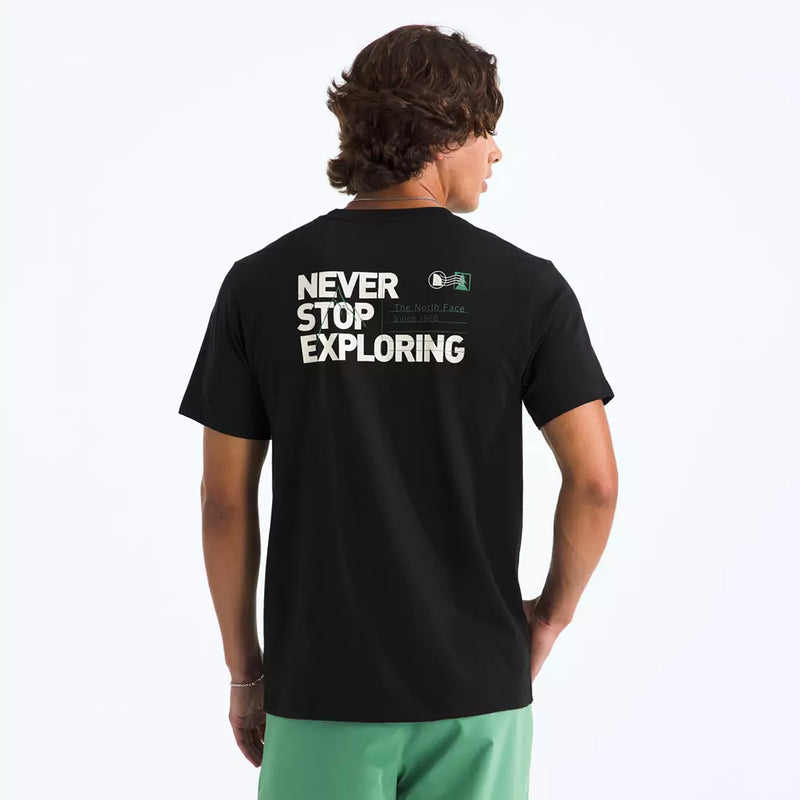 Load image into Gallery viewer, A man wearing a black The North Face t-shirt from the back, featuring the text 'NEVER STOP EXPLORING' in white, with a small green and white logo above it. He is also wearing light green shorts against a plain white background. Ski and Tennis Station.
