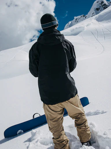 Load image into Gallery viewer, Person in black jacket and tan pants standing on a snowy mountain with a blue snowboard.
