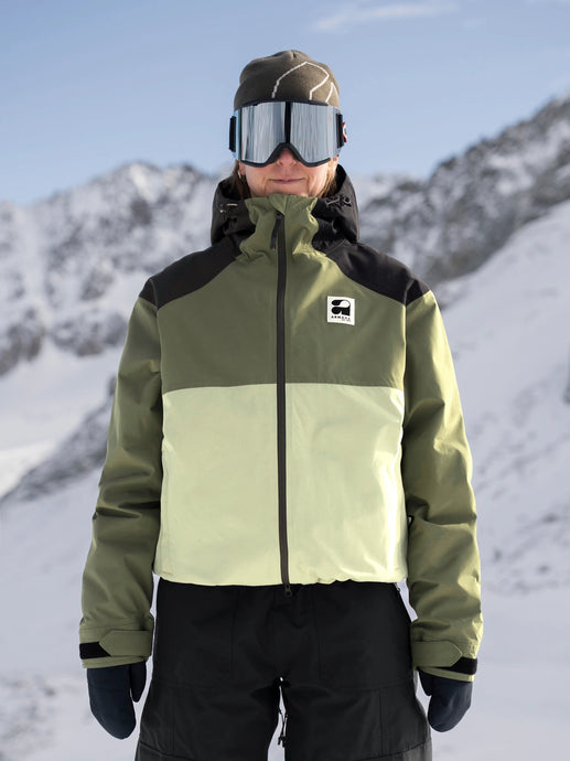 Person wearing a green and black jacket with a brand logo, standing in a snowy mountain landscape.