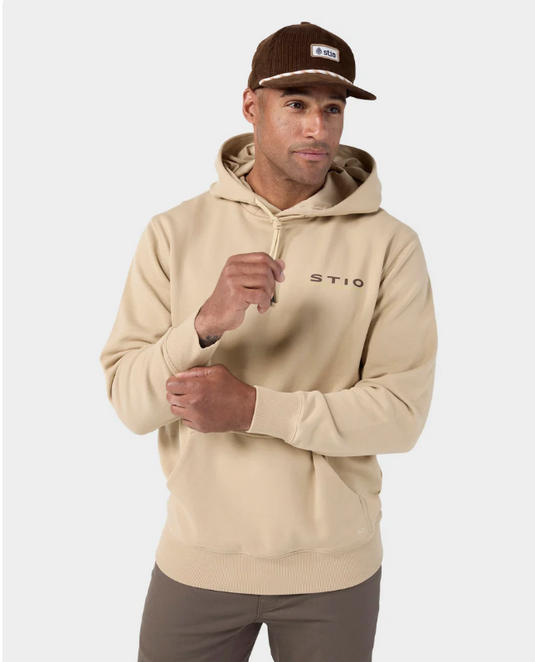 A man wearing a beige pullover hoodie with 'STIO' printed on the chest, paired with brown pants and a brown corduroy cap, against a light gray studio background. Ski and Tennis Station