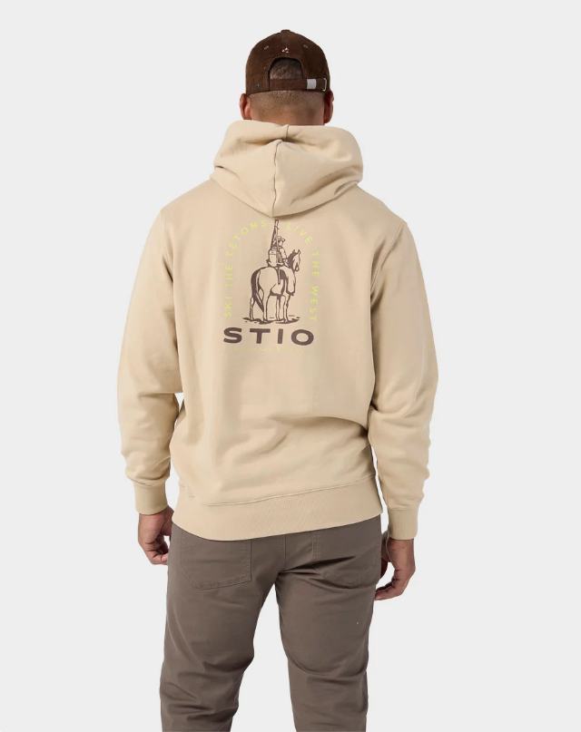 Load image into Gallery viewer, A man viewed from behind wearing a beige hoodie with a graphic of a cowboy on a horse and the text 'STIO' on the back, paired with brown pants and a brown cap. Ski and Tennis Station
