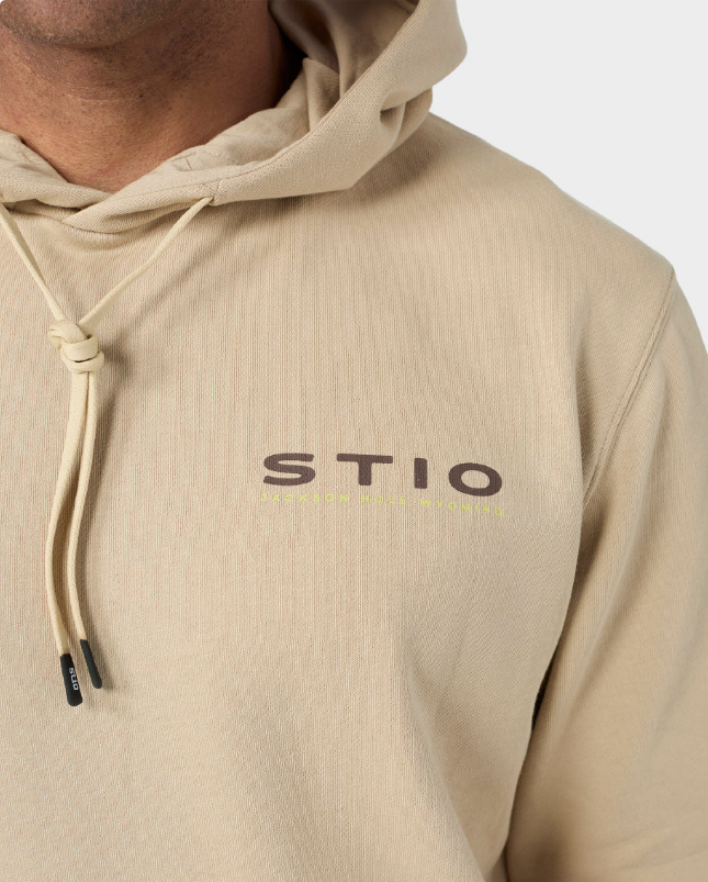Load image into Gallery viewer, Close-up of a beige hooded sweatshirt worn by a model, featuring the brand name 'STIO' printed in dark brown across the chest.
