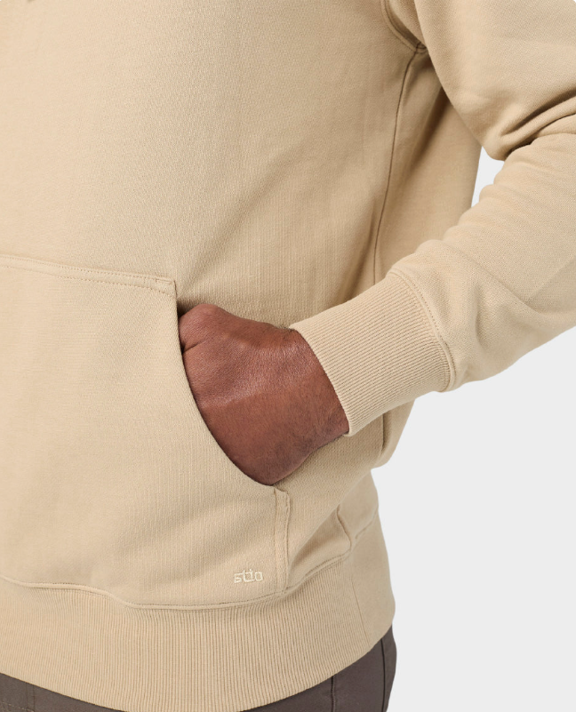 Load image into Gallery viewer, Close-up of a beige crewneck sweatshirt with a front kangaroo pocket, featuring a hand in the pocket and a small white 'stio' logo near the lower hem.
