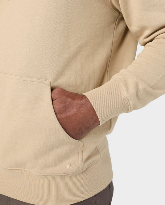 Close-up of a beige crewneck sweatshirt with a front kangaroo pocket, featuring a hand in the pocket and a small white 'stio' logo near the lower hem.