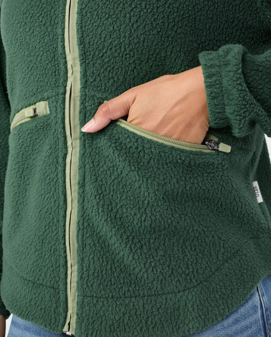 A woman modeling a medium green Stio Rawlins fleece jacket with a high collar, full-length front zipper, and two zippered hand pockets, Ski and Tennis Station