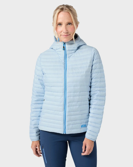 A woman modeling a light blue hooded Stio puffer jacket with a bright blue zipper, paired with navy blue leggings. Ski and Tennis Station