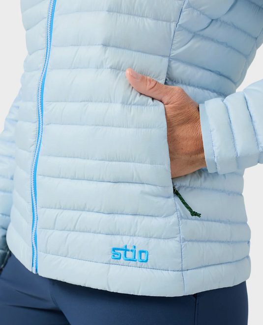 A woman modeling a light blue hooded Stio puffer jacket with a bright blue zipper, paired with navy blue leggings. Ski and Tennis Station