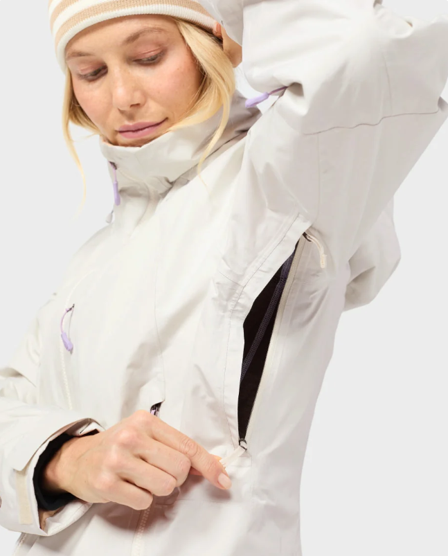 Load image into Gallery viewer, A close-up of a woman wearing a light beige technical outdoor jacket and a striped knit beanie, demonstrating a zipper pocket on the jacket.
