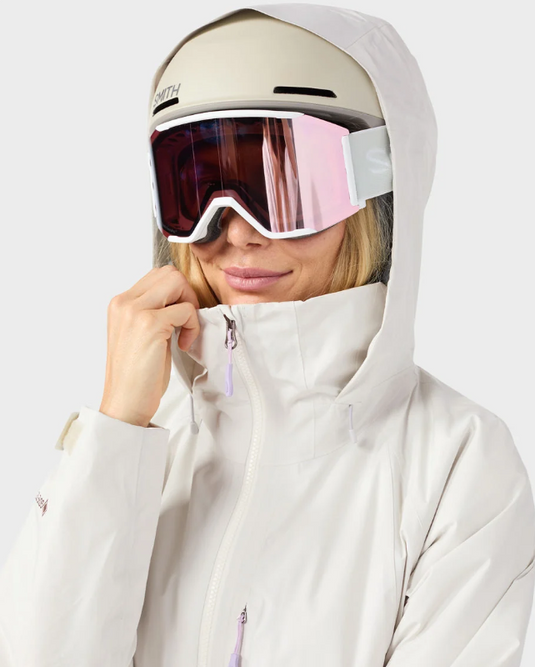 A close-up of a person wearing a light beige ski helmet, pink-tinted snow goggles, and a white ski jacket against a plain light gray background.