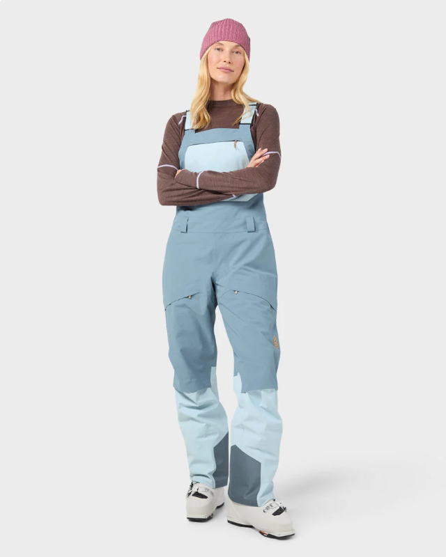 Load image into Gallery viewer, A woman modeling light blue Stio ski bib overalls with color-blocked light blue and white legs, worn over a brown long-sleeve top and a pink beanie. Ski and Tennis Station
