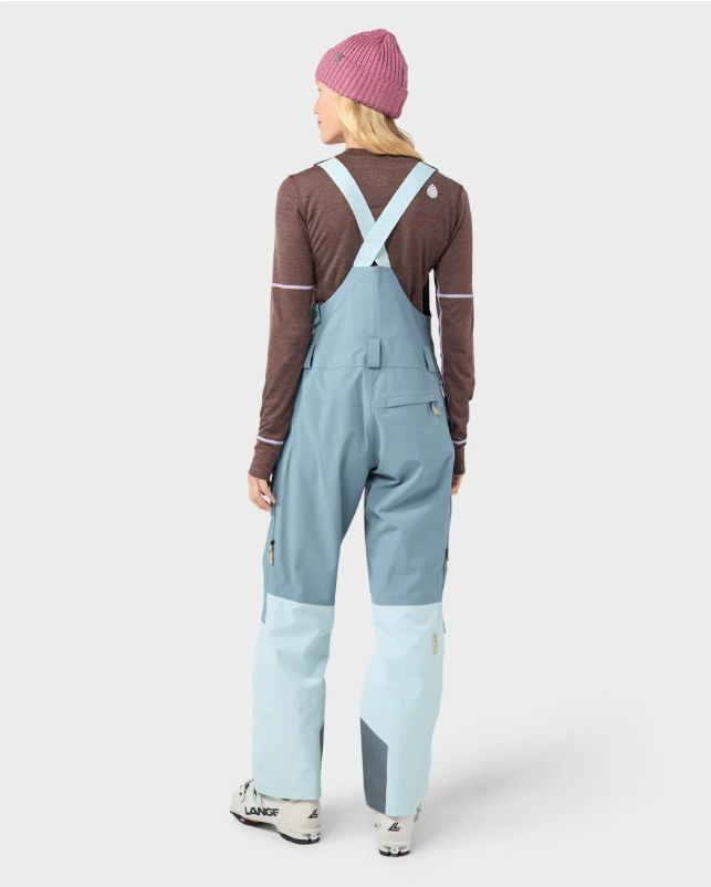 Load image into Gallery viewer, A woman modeling light blue Stio ski bib overalls with color-blocked light blue and white legs, worn over a brown long-sleeve top and a pink beanie. Ski and Tennis Station
