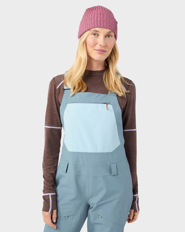 Load image into Gallery viewer, A woman modeling light blue Stio ski bib overalls with color-blocked light blue and white legs, worn over a brown long-sleeve top and a pink beanie. Ski and Tennis Station
