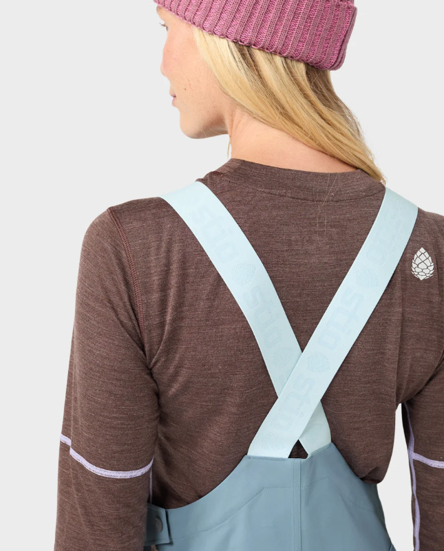 Load image into Gallery viewer, A woman modeling light blue Stio ski bib overalls with color-blocked light blue and white legs, worn over a brown long-sleeve top and a pink beanie. Ski and Tennis Station
