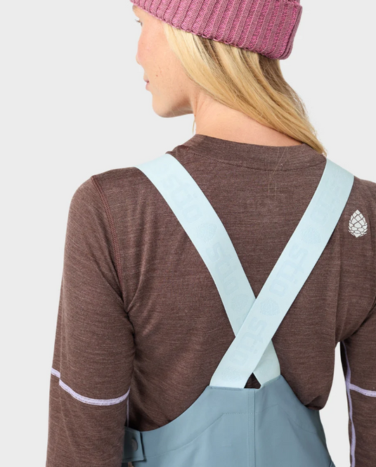 A woman modeling light blue Stio ski bib overalls with color-blocked light blue and white legs, worn over a brown long-sleeve top and a pink beanie. Ski and Tennis Station