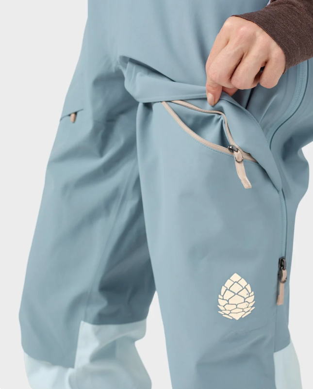 Load image into Gallery viewer, Close-up of a person wearing dusty blue ski pants with a beige drawstring waist and a white geometric pinecone logo near the lower leg.
