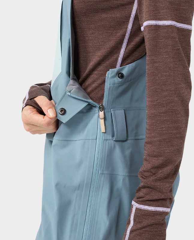 Load image into Gallery viewer, A woman modeling light blue Stio ski bib overalls with color-blocked light blue and white legs, worn over a brown long-sleeve top. Ski and Tennis Station
