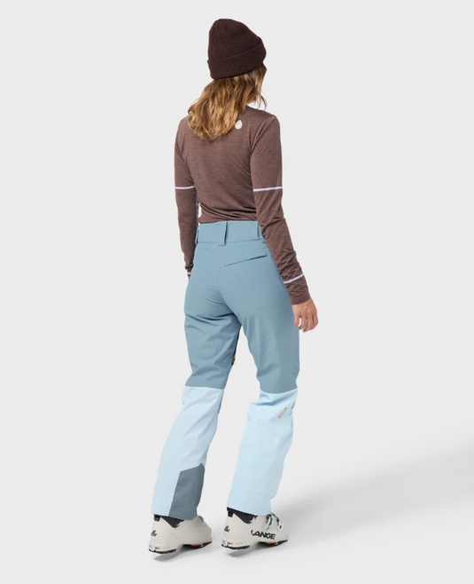A model wearing light blue and white color-blocked Stio skyrider ski pants with a brown long-sleeve top and white ski boots. Ski and Tennis Station