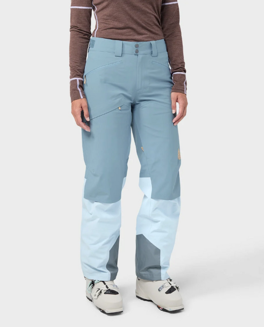 A model wearing light blue and white color-blocked Stio skyrider ski pants with a brown long-sleeve top and white ski boots. Ski and Tennis Station