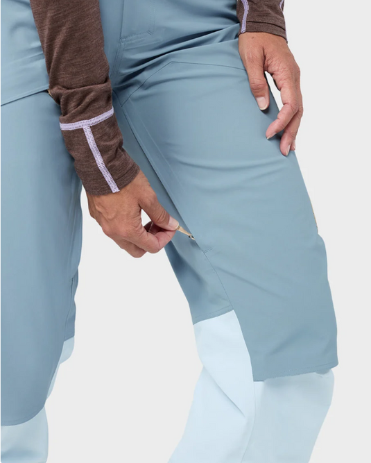 Close-up of a person wearing light blue Stio skyrider ski pants with contrasting white lower legs and a brown long-sleeve top with pale lavender trim.