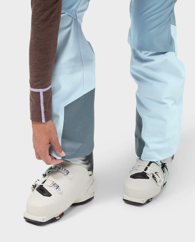 Load image into Gallery viewer, A model wearing light blue and white color-blocked Stio skyrider ski pants with a brown long-sleeve top and white ski boots. Ski and Tennis Station
