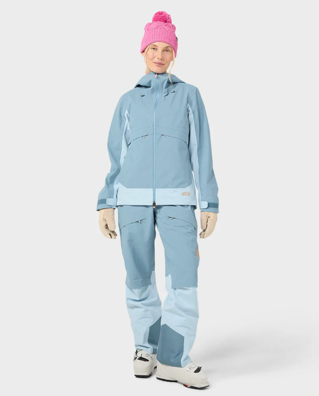 Load image into Gallery viewer, A woman wearing a light blue Stio skyrider ski jacket with contrasting pale blue panels, matching snow pants, beige gloves, and a pink knit beanie. Ski and Tennis Station
