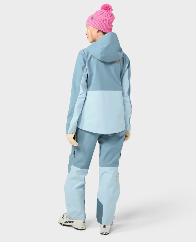 Load image into Gallery viewer, A woman wearing a light blue Stio skyrider ski jacket with contrasting pale blue panels, matching snow pants, beige gloves, and a pink knit beanie. Ski and Tennis Station
