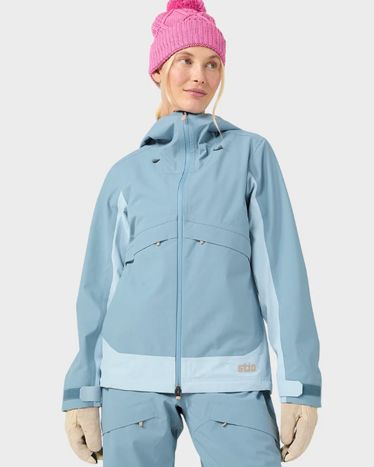 A woman wearing a light blue Stio skyrider ski jacket with contrasting pale blue panels, matching snow pants, beige gloves, and a pink knit beanie. Ski and Tennis Station