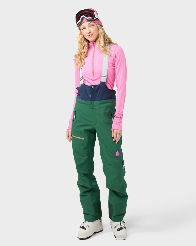 Load image into Gallery viewer, A woman modeling ski pants in forest green with a navy bib, paired with a pink long-sleeve top, a pink beanie with goggles, and white ski boots, against a light gray studio background.
