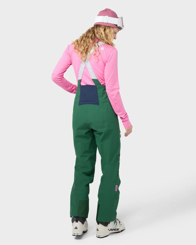 Load image into Gallery viewer, A model shown from the back wearing green ski bib overalls with a navy blue bib pocket, a pink long-sleeve shirt, a pink and white striped beanie, and ski boots against a light gray studio background.
