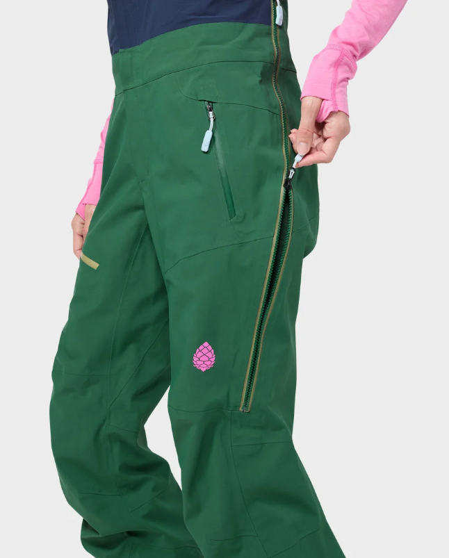 Load image into Gallery viewer, Close-up of a person wearing dark green stio ski pants with a pink long-sleeve top, showing the side pocket, zipper, and a small pink logo patch on the leg against a light gray background.
