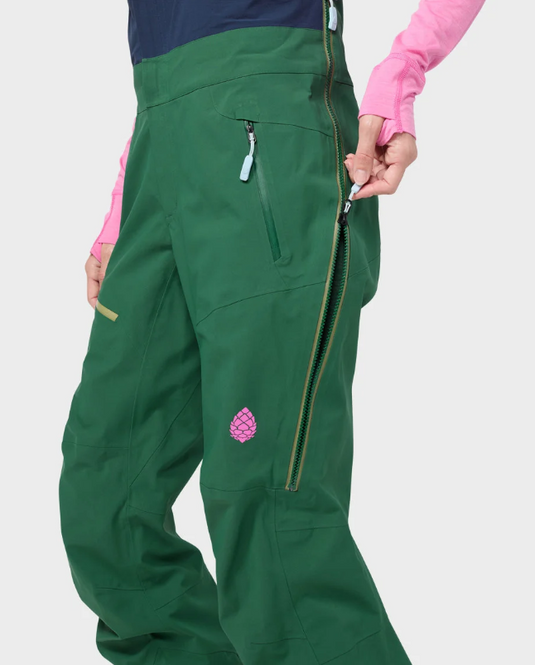 Close-up of a person wearing dark green stio ski pants with a pink long-sleeve top, showing the side pocket, zipper, and a small pink logo patch on the leg against a light gray background.