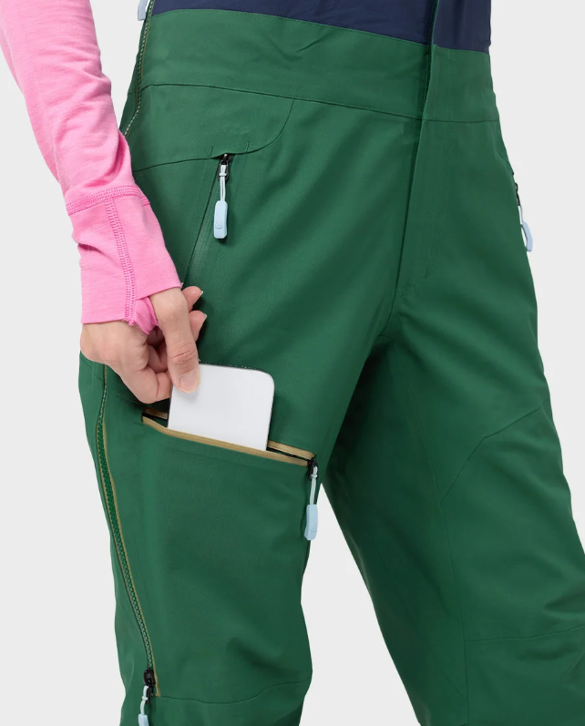 Load image into Gallery viewer, Close-up of a person wearing green ski pants with a pink long-sleeve top, showing a side pocket holding a white phone.
