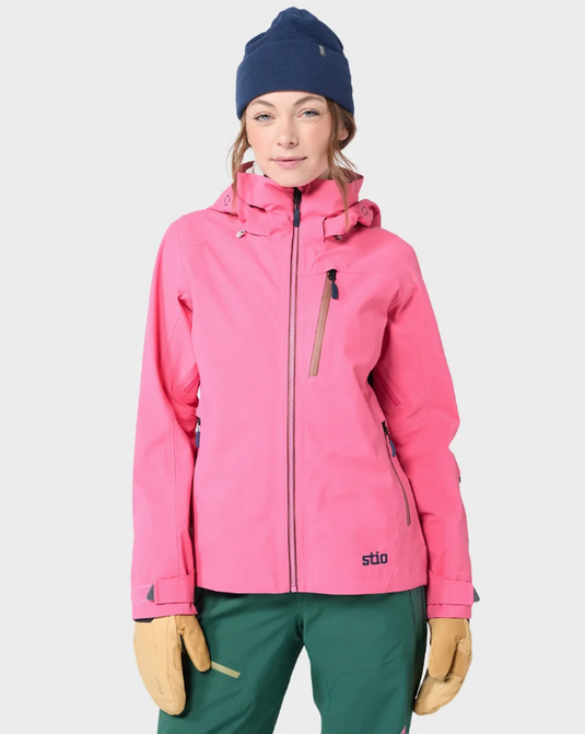 A woman modeling a bright pink ski jacket with a navy beanie, green pants, and tan gloves against a light gray studio background. The brand name 'stio' is visible on the lower left of the jacket. Ski and Tennis Station