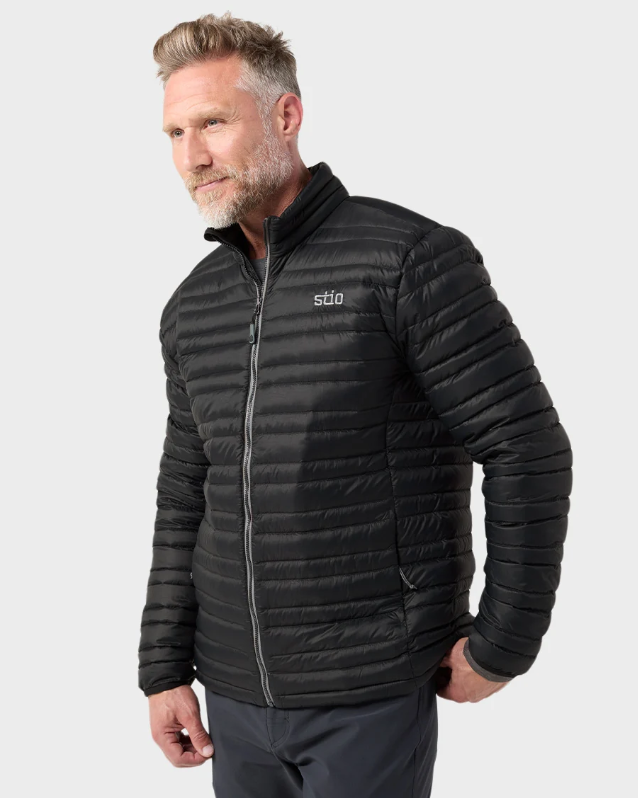 Load image into Gallery viewer, A man wearing a black Stio puffer jacket with horizontal quilting and a silver zipper, shown from the waist up against a light gray studio background. The jacket has a small white 'stio' logo on the left chest. Ski and Tennis Station
