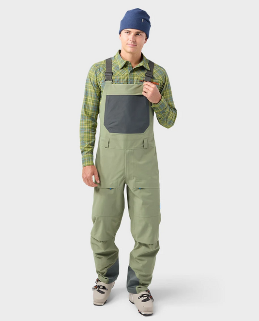 A man modeling olive green Stio bib overalls with a dark gray chest pocket, worn over a green and yellow plaid shirt. Ski and Tennis Station.