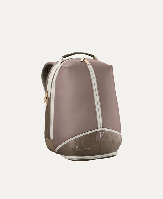 A minimalist Roger Federer Hall of Fame backpack in a muted taupe-brown color with cream-colored piping and gold-tone zipper pulls, shown from a three-quarter angle against a solid off-white background. Special Edition. Ski and Tennis Station.
