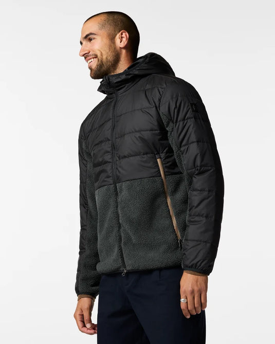 A man wearing a black Cotopaxi hooded puffer jacket, the Trico 2.0, with a dark gray fleece lower panel and a brown zipper accent, paired with dark pants, photographed against a clean white studio background. Ski and Tennis Station