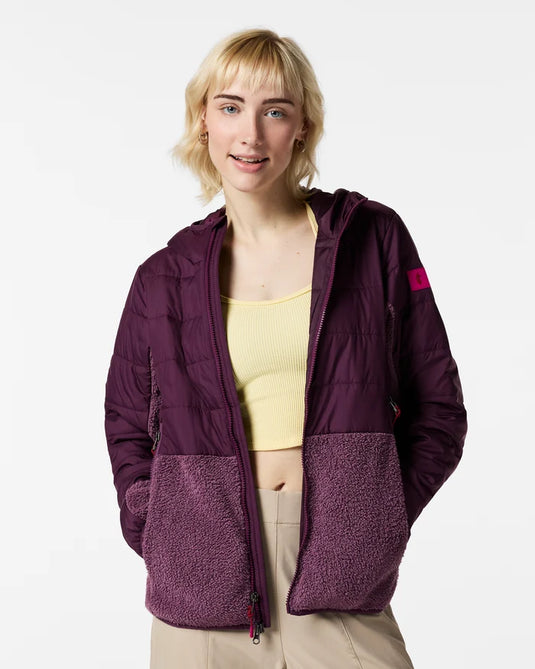 A woman modeling a dark purple hooded Cotopaxi Trico 2.0 hybrid puffer jacket with a textured fleece lower panel and a small pink logo patch on the sleeve, worn over a pale yellow ribbed tank top and beige pants. Ski and Tennis Station